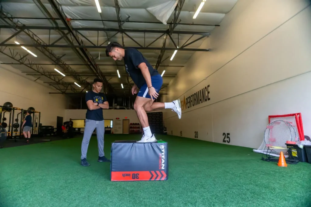 Athlete performing a plyometric box jump with coach supervision in a gym